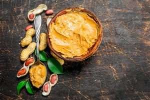 A wooden bowl of peanut butter with peanuts and a knife on a dark wooden table