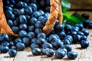 A pile of fresh blueberries spilling out of a woven basket onto a wooden surface with green leaves scattered around
