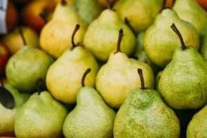 A pile of green and yellow pears with brown stems, in front of a blurred background of other fruits