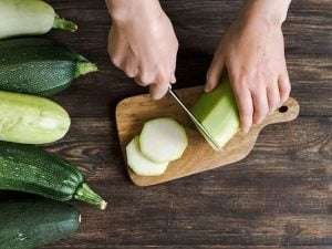 Person slicing light green zucchini on wooden cutting board with silver knife and black handle, surrounded by dark green zucchinis of varying sizes