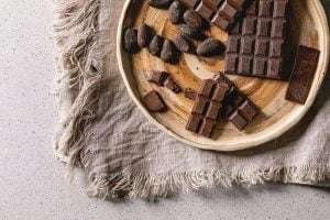A wooden plate with dark chocolate and cocoa beans on a textured tablecloth