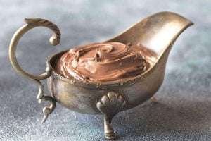 A vintage brass gravy boat with a sea horse handle and shell spout filled with chocolate mousse on a blue-grey surface
