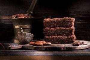 Three slices of chocolate cake on a wooden cutting board with cocoa beans and a silver teapot