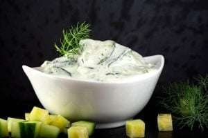 A white, modern, angular bowl of creamy white tzatziki sauce with green flecks of dill and cucumber on a black textured background