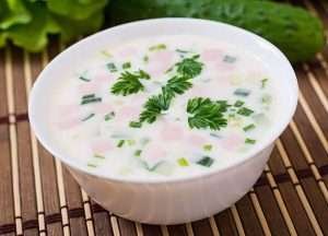 A white bowl of cold soup with pink meat and green herbs on a bamboo mat