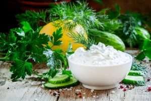 A white bowl of Yogurt on a rustic wooden table surrounded by herbs and vegetables such as dill, parsley, cucumber, lemon, and pink peppercorns