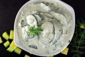 White bowl of tzatziki sauce with herbs and sliced cucumbers on a black background
