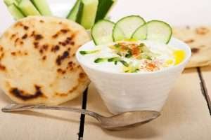 A bowl of tzatziki sauce with pita bread and sliced cucumbers on a wooden table