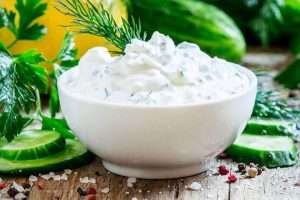 White bowl of tzatziki sauce on a wooden table with scattered herbs, cucumber slices, and pink peppercorns