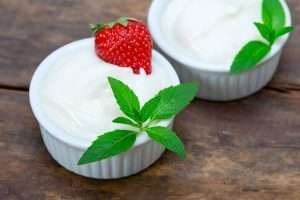 Two small white ramekins of vanilla yogurt with mint and strawberry on a wooden surface