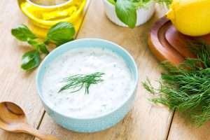 A blue bowl of tzatziki sauce with green herbs on a wooden cutting board with lemon, basil, dill, and olive oil