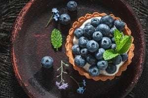 A blueberry tart with a golden crust, white cream, and fresh blueberries on a red plate with mint and purple flowers on a dark woven mat