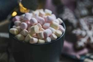 A black mug filled with pink and white marshmallows on a table with a pink and white checkered tablecloth and string lights in the background
