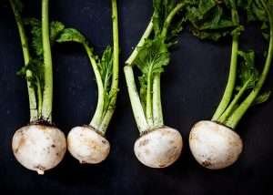 Four freshly harvested turnips with green stems and leaves on a black platter