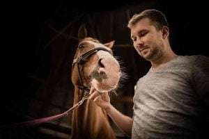 A man with a blurred face standing next to a brown horse in a stable